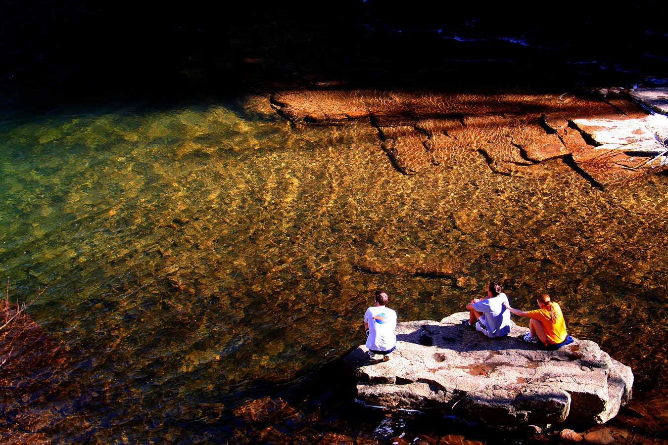 Viewed from above, three people sit on a rock surrounded by rippled, see-through water that varies in color from roange to green.