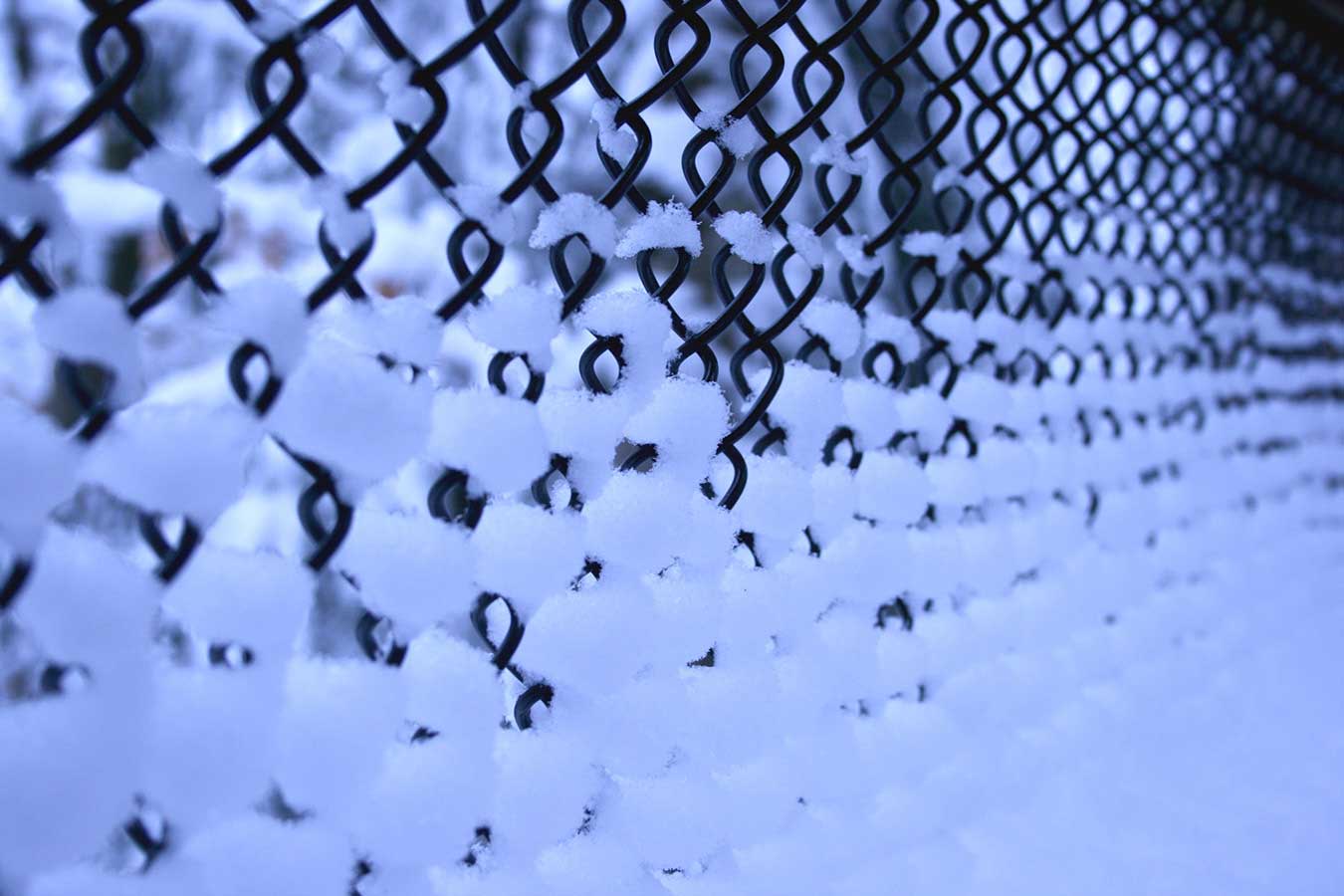 A chain link fence fills the frame and is angled away to the right. The rear is out of focus. Snow puffs fill each chain link diamond, increasing in volume from the top to the bottom of the frame.